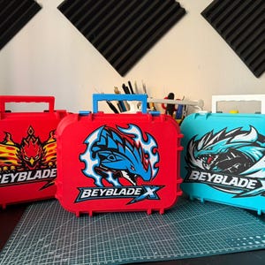 May include: Three colorful Beyblade X storage cases. The cases are red and teal, with graphic designs and the Beyblade X logo. Each case has a handle for carrying.