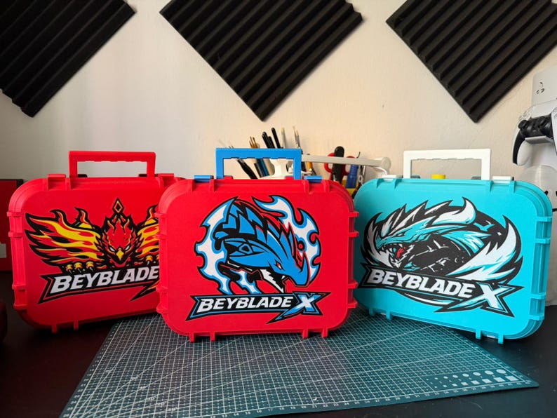 May include: Three colorful Beyblade X storage cases. The cases are red and teal, with graphic designs and the Beyblade X logo. Each case has a handle for carrying.