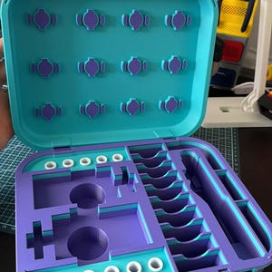 May include: An open, two-tone organizer case in teal and purple. The top lid has several purple, decorative accents. The interior features various compartments and slots for storage. The case has a handle and is designed for organization.