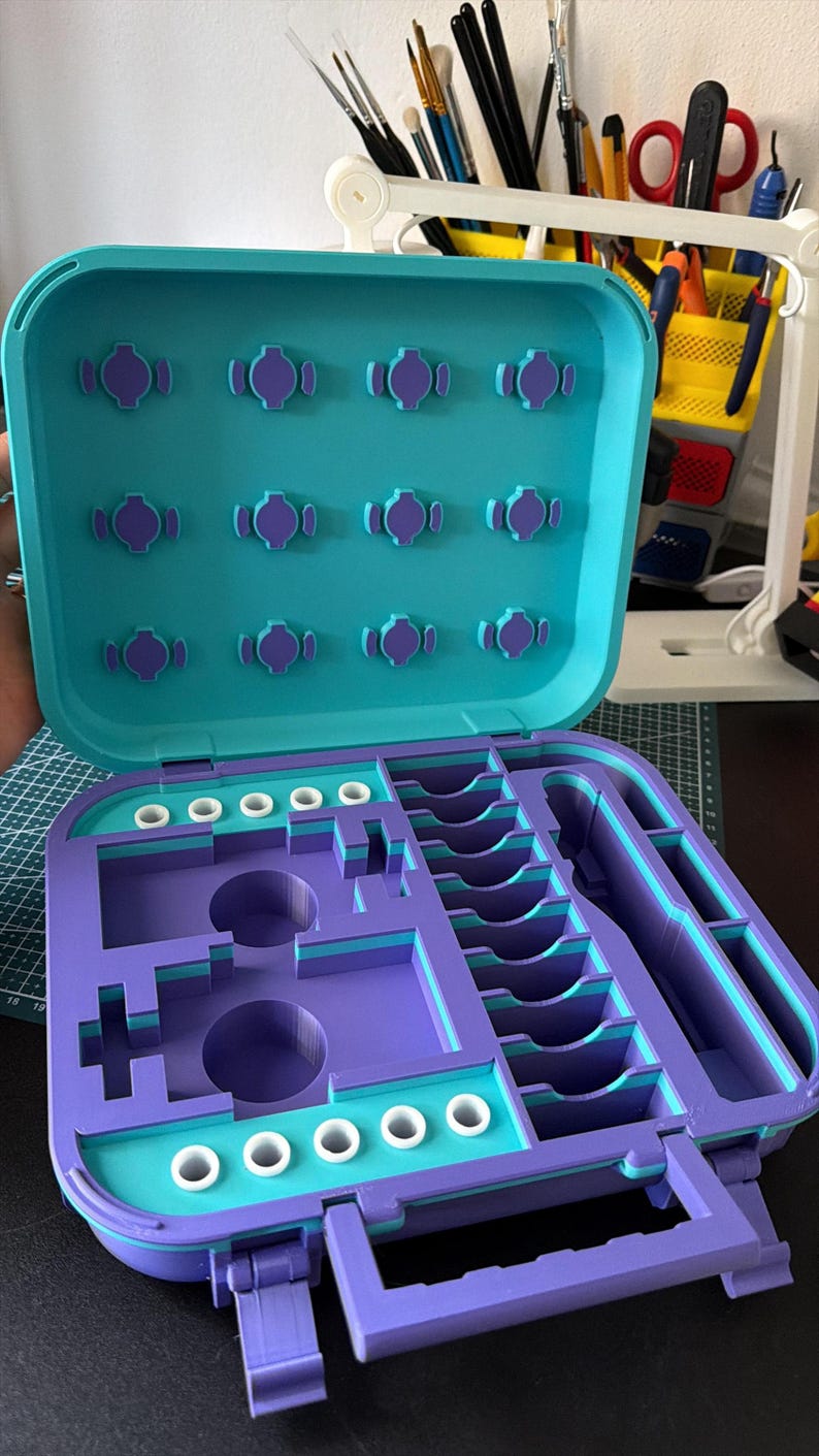 May include: An open, two-tone organizer case in teal and purple. The top lid has several purple, decorative accents. The interior features various compartments and slots for storage. The case has a handle and is designed for organization.