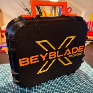 May include: Black case with an orange handle and accents. The case features the text "BEYBLADE" in orange with a stylized "X" and the word "PEODORO" below. The case is sitting on a green cutting mat.