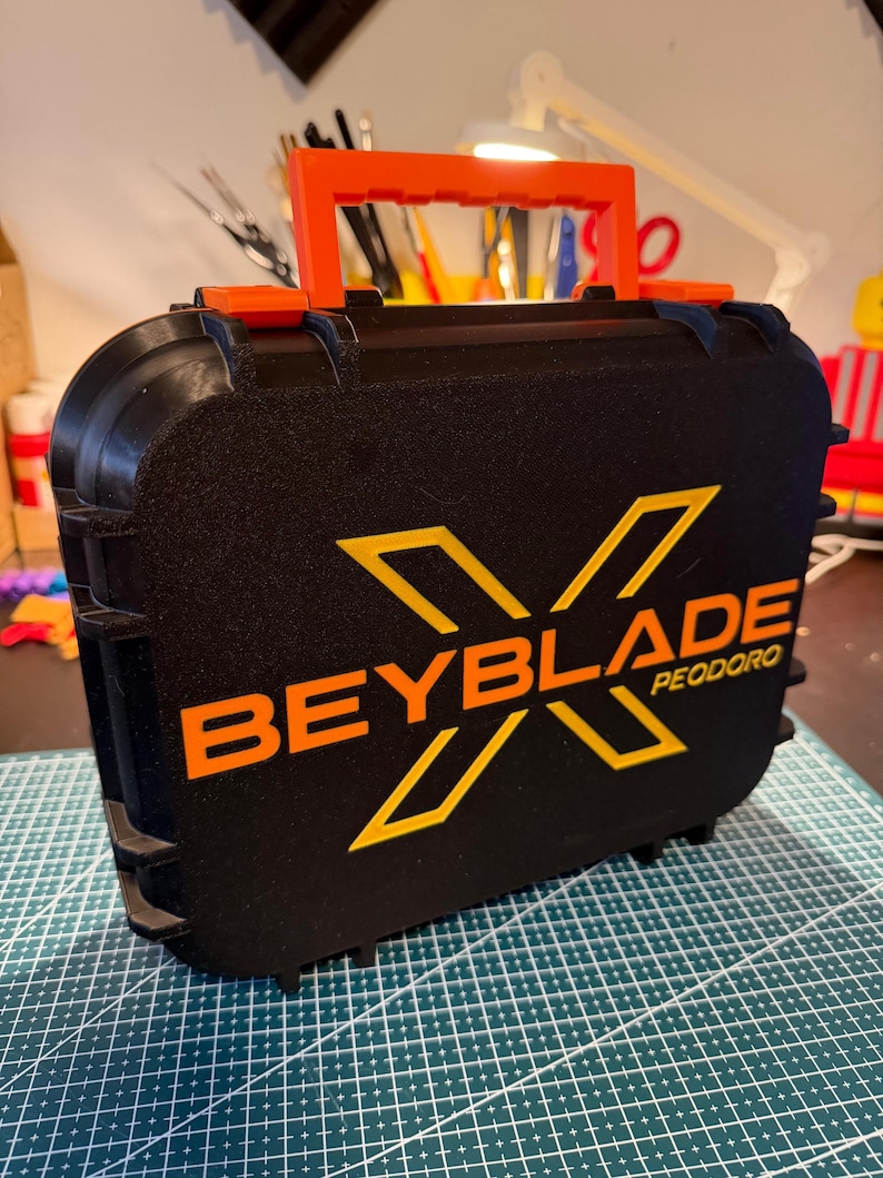 May include: Black case with an orange handle and accents. The case features the text "BEYBLADE" in orange with a stylized "X" and the word "PEODORO" below. The case is sitting on a green cutting mat.