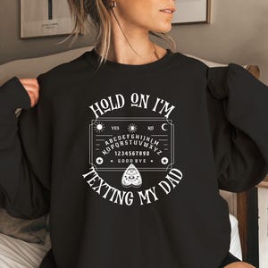 May include: A black long-sleeve sweatshirt featuring a white graphic print of a Ouija board design. The text reads "HOLD ON I'M TEXTING MY DAD" in a curved font above and below the board, which includes the alphabet, numbers, and a planchette.
