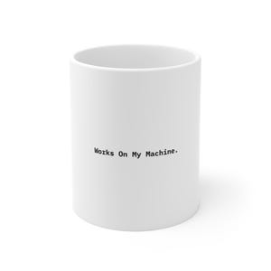 May include: A white ceramic mug featuring the black text Works On My Machine. printed in a simple sans-serif font on the front. The mug has a cylindrical shape with a smooth finish and a wide opening at the top, standing upright against a plain background.