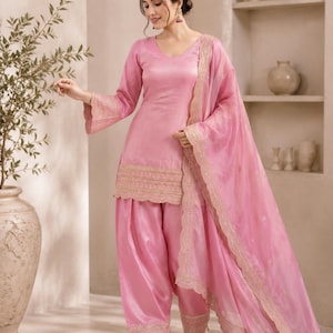 Könnte beinhalten: Ein hellrosa dreiteiliges Outfit bestehend aus einer Tunika mit V-Ausschnitt und langen Ärmeln, einer passenden locker sitzenden Hose und einem transparenten Dupatta. Tunika und Hose haben goldene, bestickte Bogenkanten, Logo Leverice.
