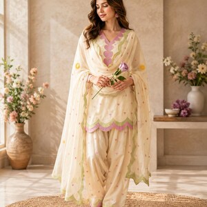 Op de afbeelding: Een crèmekleurige salwar kameez-set bestaande uit een tuniek met een geschulpte zoom, een bijpassende broek en een transparante dupatta. Het kledingstuk is versierd met kleine roze hartjespatronen en groene geschulpte randen.