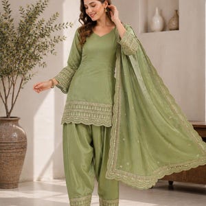Könnte beinhalten: Ein salbeigrünes Salwar-Kameez-Set bestehend aus einer Tunika mit V-Ausschnitt und dreiviertellangen Glockenärmeln, einer passenden Plisseehose und einem transparenten Dupatta. Saum, Ärmel und Hose sind mit goldener Stickerei verziert.