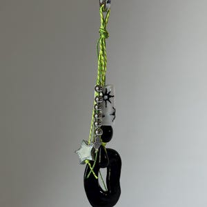 May include: A keychain featuring a neon green cord attached to a silver metal clip. The cord holds a white cylindrical bead with black star patterns, several small silver spherical beads, a small silver hamsa charm, a white star charm, and a large black irregular oval-shaped pendant.