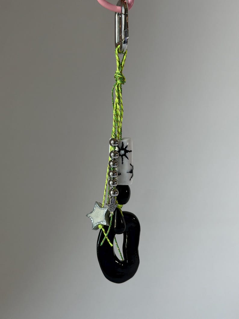 May include: A keychain featuring a neon green cord attached to a silver metal clip. The cord holds a white cylindrical bead with black star patterns, several small silver spherical beads, a small silver hamsa charm, a white star charm, and a large black irregular oval-shaped pendant.