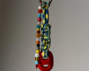 May include: A keychain featuring a silver-toned metal clasp attached to a braided blue and yellow paracord. A single strand of multicolored beads in red, blue, yellow, white, orange, and grey hangs alongside a solid, glossy red plastic ring.