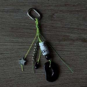 May include: A keychain featuring a silver-toned metal carabiner attached to neon green strings. The strings hold a grey star charm, a strand of small silver beads, a white cylindrical bead with black sun motifs, and a large black organic-shaped pendant.