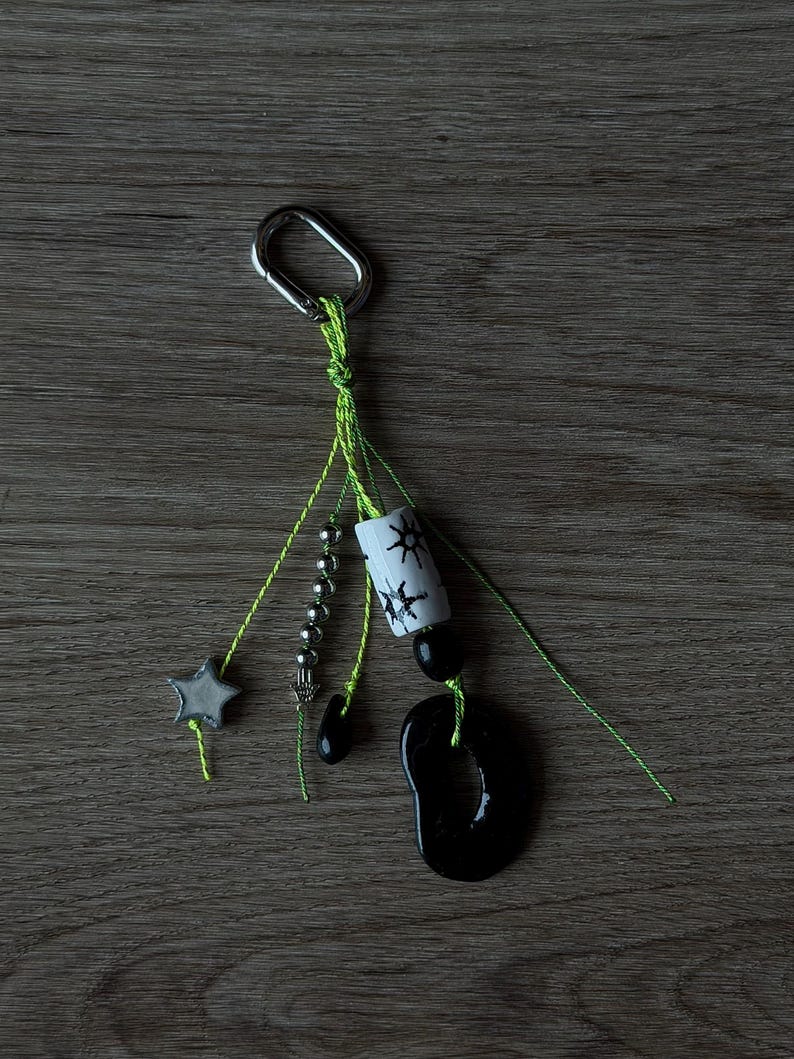 May include: A keychain featuring a silver-toned metal carabiner attached to neon green strings. The strings hold a grey star charm, a strand of small silver beads, a white cylindrical bead with black sun motifs, and a large black organic-shaped pendant.