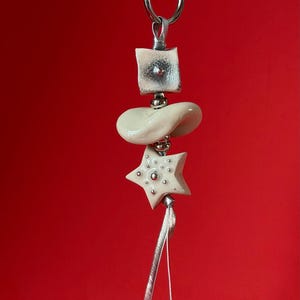 May include: A decorative hanging ornament featuring a silver-toned metal ring at the top, followed by a square metallic bead, a smooth cream-colored curved ceramic piece, and a cream-colored star-shaped bead with small silver dots, ending in a thin ribbon.
