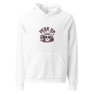 Peut inclure: Un sweat à capuche blanc avec une poche avant et une capuche à cordon. Le graphisme sur le devant représente une tasse de café souriante avec les mots "PERK UP" au-dessus. Des grains de café et une machine à café sont également dans le graphisme.