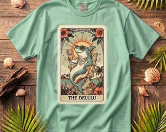 May include: A pale green t-shirt with a tarot card graphic. The card depicts a shark in sunglasses and a necklace, with the text "THE DELULU". The design includes floral and tropical imagery.