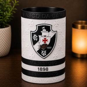 May include: A cylindrical black and white ceramic mug with the Vasco da Gama crest. The mug has the words "VASCO DA GAMA", "MAJESTOSO", "1898", "&Eacute; SENTIMENTO", and a repeating pattern of crosses. The mug is on a wooden surface.
