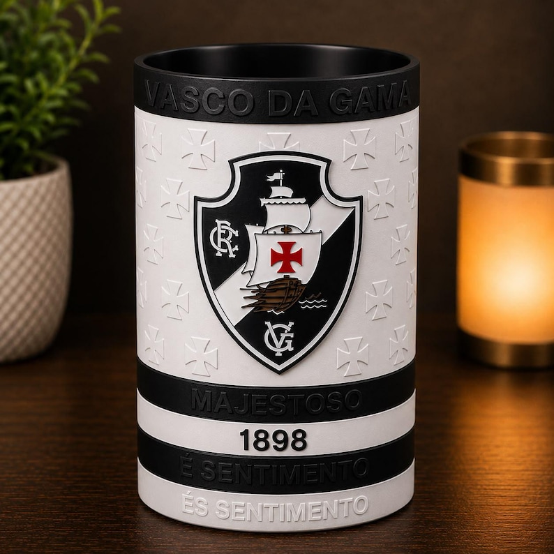 May include: A cylindrical black and white ceramic mug with the Vasco da Gama crest. The mug has the words "VASCO DA GAMA", "MAJESTOSO", "1898", "&Eacute; SENTIMENTO", and a repeating pattern of crosses. The mug is on a wooden surface.