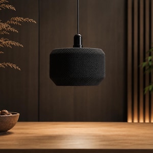 May include: A black cylindrical pendant light fixture with a fine, textured mesh surface. The lamp hangs from a thin black cord and features a smooth, solid black cap at the top where the cord connects to the main body of the light fixture.