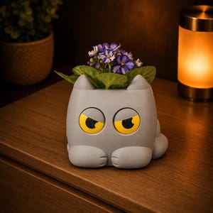 May include: A gray cat-shaped planter with yellow eyes and a purple flower arrangement. The planter has a whimsical design, with a cat face and paws. The flowers are a vibrant contrast to the gray planter.