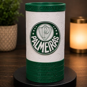 May include: A white and green cylindrical tumbler with the Palmeiras logo. The design includes the team's emblem, the words "PARQUE CANTA E VIBRA", and "SOCIEDADE ESPORTIVA PALMEIRAS DESDE 1914". The tumbler is on a black base.