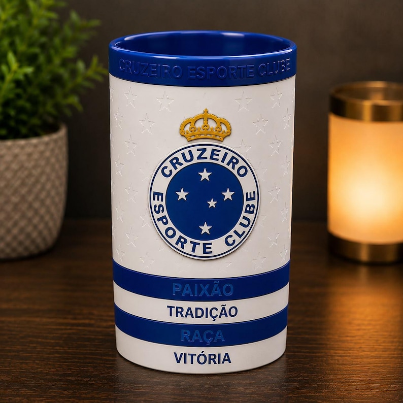 May include: A white and blue Cruzeiro Esporte Clube mug. The mug features the team's logo, a gold crown, and the words "PAIX&Atilde;O, TRADI&Ccedil;&Atilde;O, RA&Ccedil;A, VIT&Oacute;RIA". The top and interior are blue. The mug is decorated with star patterns.