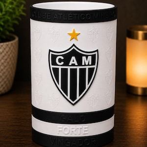 May include: White cylindrical tumbler with the "CAM" logo in black and white, and a gold star. The design includes the words "CLUBE ATL&Eacute;TICO MINEIRO", "GALO FORTE", and "VINGADOR" in black. The tumbler has black bands at the top and bottom.
