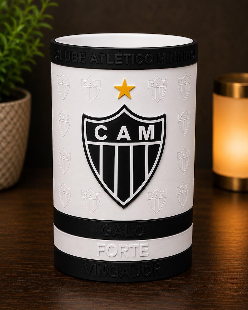 May include: White cylindrical tumbler with the "CAM" logo in black and white, and a gold star. The design includes the words "CLUBE ATL&Eacute;TICO MINEIRO", "GALO FORTE", and "VINGADOR" in black. The tumbler has black bands at the top and bottom.