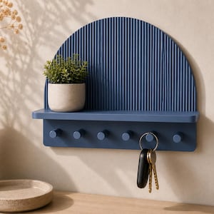 May include: A wall-mounted navy blue shelf featuring a semi-circular back panel with a vertical ribbed texture. The shelf includes a flat surface for small items and a lower horizontal bar equipped with six cylindrical pegs for hanging keys.