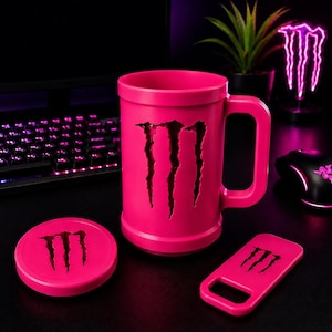 May include: A bright pink mug with a large handle, featuring a cutout of the Monster Energy logo on the front. Included are a matching pink circular coaster and a rectangular bottle opener, both displaying the black Monster Energy logo.