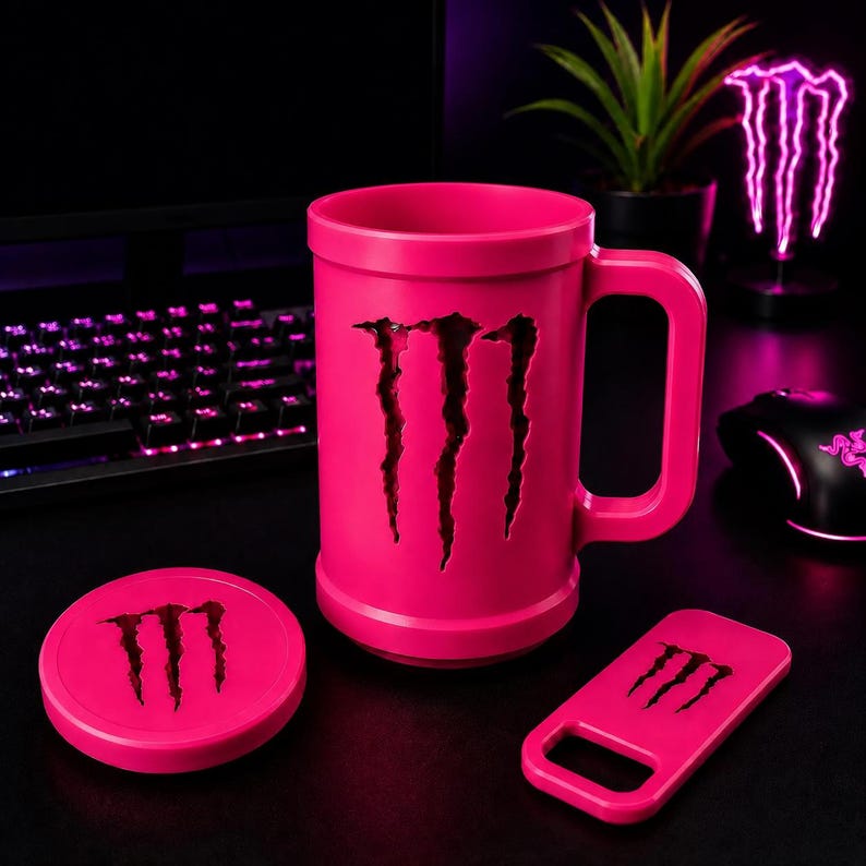 May include: A bright pink mug with a large handle, featuring a cutout of the Monster Energy logo on the front. Included are a matching pink circular coaster and a rectangular bottle opener, both displaying the black Monster Energy logo.