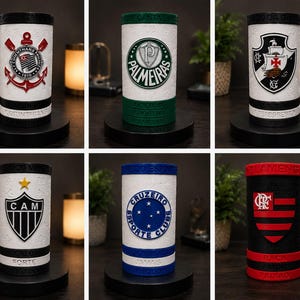 May include: Cylindrical drink holders with team logos and colours. The holders are white with black accents and sit on black bases. Logos include a cross, a star, and a shield. Each has text at the base.