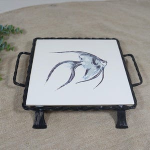 Può includere: Una piastrella quadrata in ceramica bianca con un'illustrazione stilizzata di un pesce angelo grigio e nero, inserita in una cornice di metallo nero con manici laterali intrecciati e quattro gambe corte come base decorativa.