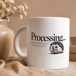 May include: A white ceramic mug featuring a black graphic of a hedgehog and the text "Processing... MORE DRAMATICALLY THAN NECESSARY" printed on the front. The mug has a standard handle and a smooth, glossy finish, displaying a clear, dark font.