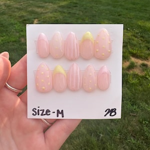 May include: A set of medium-sized, oval-shaped press-on nails. The nails are primarily light pink with yellow polka dots and accents. The text "Size-M" and "2B" are printed on the white card.