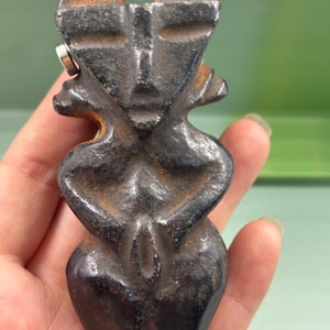 May include: A dark, weathered, cast iron figurine featuring a stylized human form with a flat, angular face, prominent brow, and arms folded across the torso. The surface shows signs of oxidation and rust, with a small silver-colored metal pin.