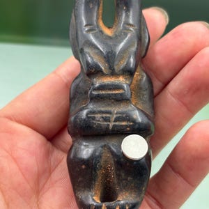 Puede incluir: Una figura negra tallada con la forma de un animal estilizado, posiblemente un conejo o una liebre, con orejas prominentes y una postura sentada. La figura tiene un aspecto envejecido con textura visible y un pequeño objeto redondo plateado.