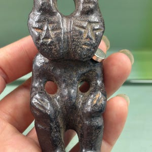 May include: A dark brown, carved stone figurine featuring two conjoined faces at the top and a seated figure below. The piece has a smooth, weathered texture with visible circular cutouts between the arms and legs, and a hole at the very top.