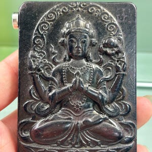 May include: A rectangular black plaque featuring a relief carving of a seated deity with four arms. The figure is depicted in a meditative pose with hands joined at the chest, surrounded by an ornate halo and floral motifs on a dark surface.