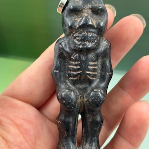 May include: A small, dark gray figurine of a skeletal figure with a detailed rib cage and skull-like face. The figurine has a small silver ring attached to the top of its head. The figure is held in a hand against a blurred green background.