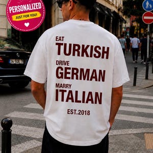 May include: A white short-sleeved t-shirt featuring dark red text on the back that reads EAT TURKISH, DRIVE GERMAN, MARRY ITALIAN, and EST. 2018. The shirt has a crew neckline and a relaxed fit, with the text printed in a bold, sans-serif font.