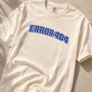 Puede incluir: Una camiseta de manga corta de color crema con el texto ERROR:404 impreso en una fuente azul llamativa de estilo universitario en el pecho. La prenda tiene cuello redondo y una etiqueta de marca visible en el interior del cuello que indica la talla L.