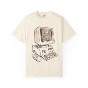 Peut inclure: Un t-shirt à manches courtes de couleur crème avec une illustration graphique d'un ordinateur vintage. L'écran affiche le texte FAILED BOOT, la base de l'ordinateur porte les lettres FC, et une souris d'ordinateur est connectée à l'appareil.