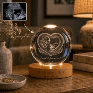 May include: A spherical glass ornament featuring an etched ultrasound image inside a heart shape, with the text "Loved before we met" and "2026". It sits on a round wooden base engraved with "To My Baby : Hunt" and "June 2026".