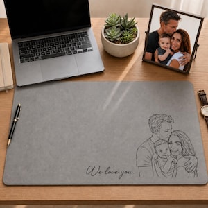 Könnte beinhalten: Eine graue Schreibtischunterlage mit einer schwarzen Strichzeichnung einer Familie, bestehend aus einem Elternteil, einem Kind und einem Baby, mit dem Text "We love you." in Schreibschrift. Die Unterlage liegt auf einer Holzoberfläche.