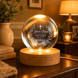 Può includere: Una sfera di vetro trasparente incisa con una croce, un fonte battesimale e il testo James Tatum BAPTIZED Sep 12, 2026, poggiata su una base rotonda in legno con l'iscrizione Wonderfully made, deeply loved, and forever a precious child of God.