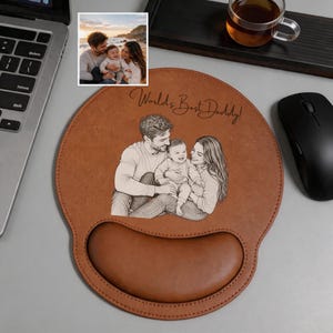 May include: A tan leather mouse pad with a wrist rest featuring a laser-engraved illustration of a family of three. The text World's Best Daddy! is engraved above the illustration, which depicts a man, a woman, and a baby sitting together.