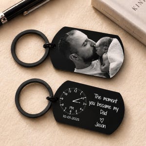 May include: Two black metal keychains. One features a black and white photo of a man kissing a baby. The other keychain has a clock design with the text "The moment you became my Dad" and the name "Jason".