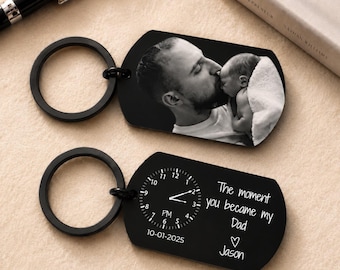 Pode incluir: Dois chaveiros de metal preto. Um apresenta uma foto em preto e branco de um homem beijando um bebê. O outro chaveiro tem um design de relógio com o texto "The moment you became my Dad" e o nome "Jason".