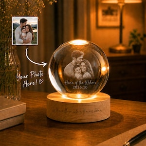 Peut inclure: Une lampe sphérique en verre avec une image gravée au laser d'une famille avec un enfant, posée sur une base cylindrique en bois. La base porte l'inscription 'Where there is love, there is home' et le verre affiche '2026.10'.