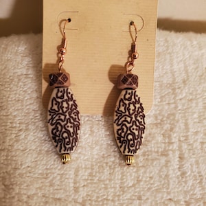 May include: A pair of dangle earrings featuring copper-toned hooks, small square brown beads with etched patterns, elongated oval cream-coloured beads with dark brown organic line designs, and tiny metallic gold-toned beads at the bottom tips.